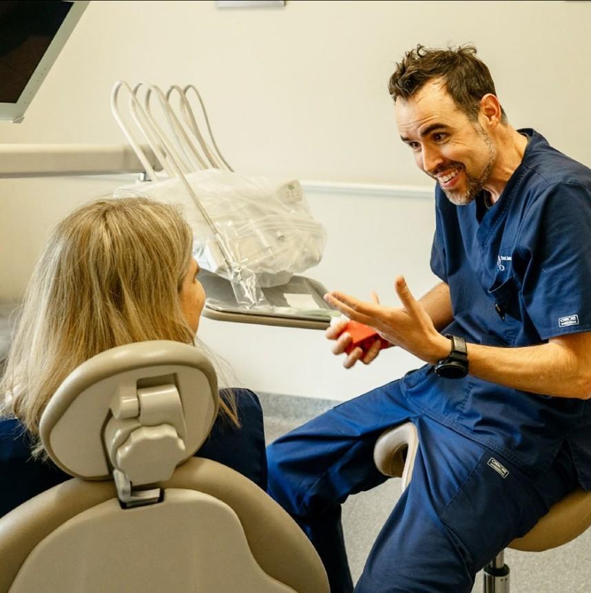 Dr. Andre Schertel in blue scrubs consults with a patient in a dental chair, gesturing while holding a small red object.